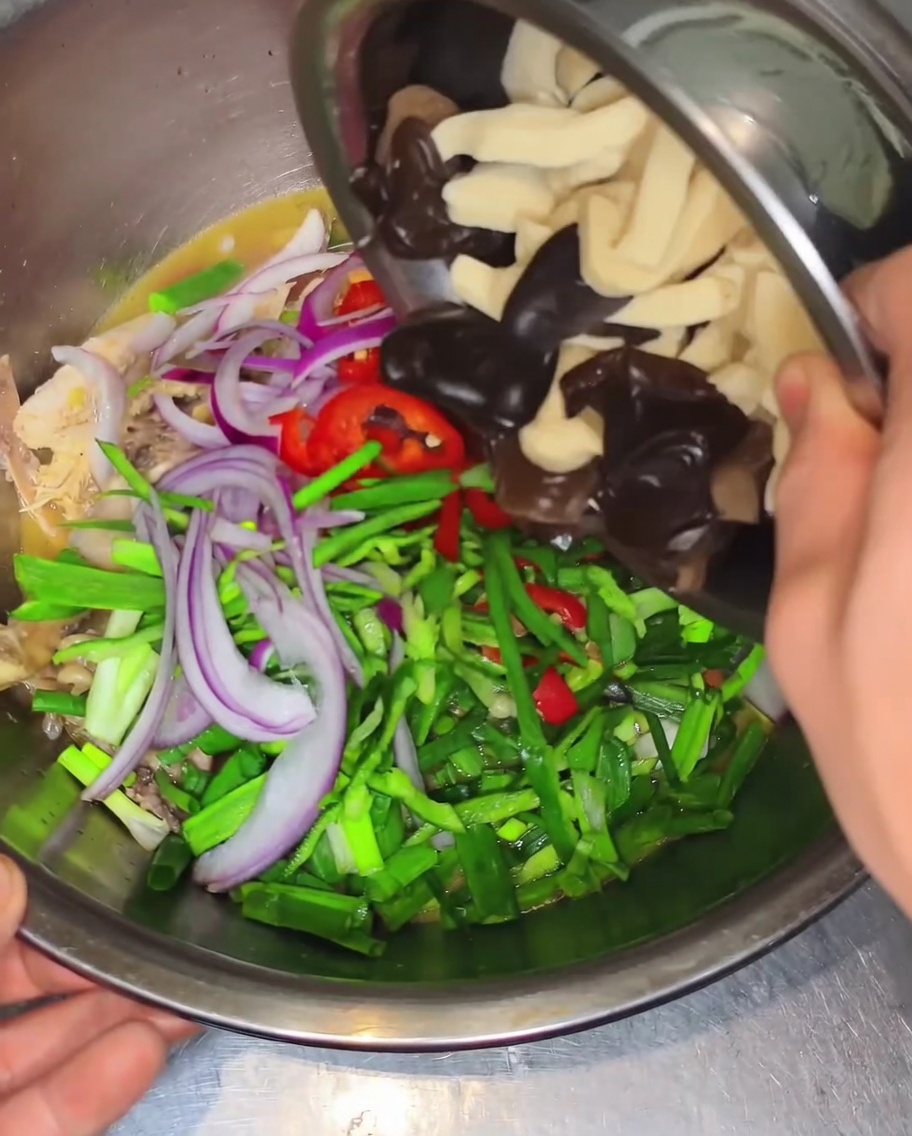 Hands tipping a small metal bowl to add dark wood ear mushrooms and thick tofu skin slices into a large mixing bowl filled with chicken, peppers, and onions.
