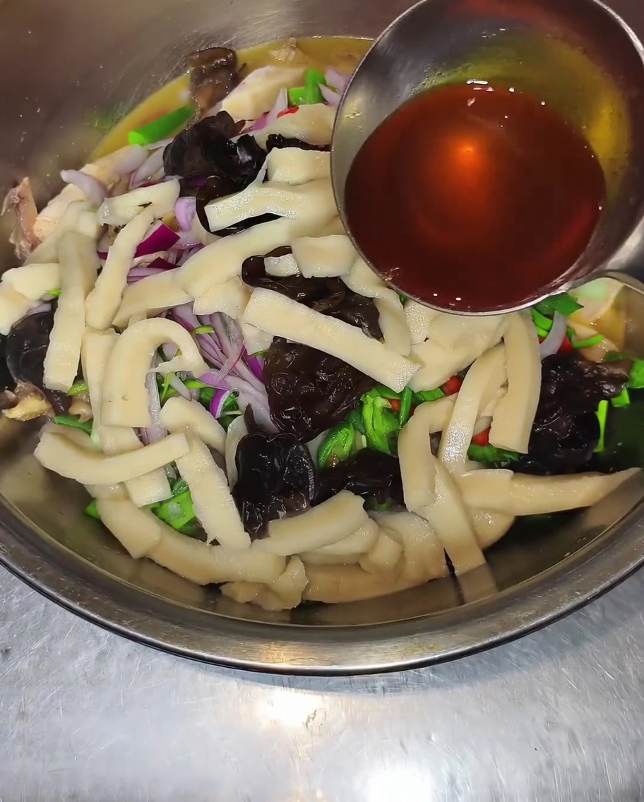 A ladle drizzling dark chili and peppercorn oil into a bowl containing chicken, wood ear mushrooms, sliced onions, and green peppers.