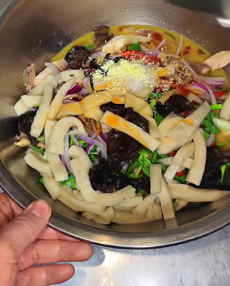 A close-up of a bowl filled with chicken, bamboo shoots, wood ear mushrooms, and peppers being seasoned with chicken bouillon and salt.