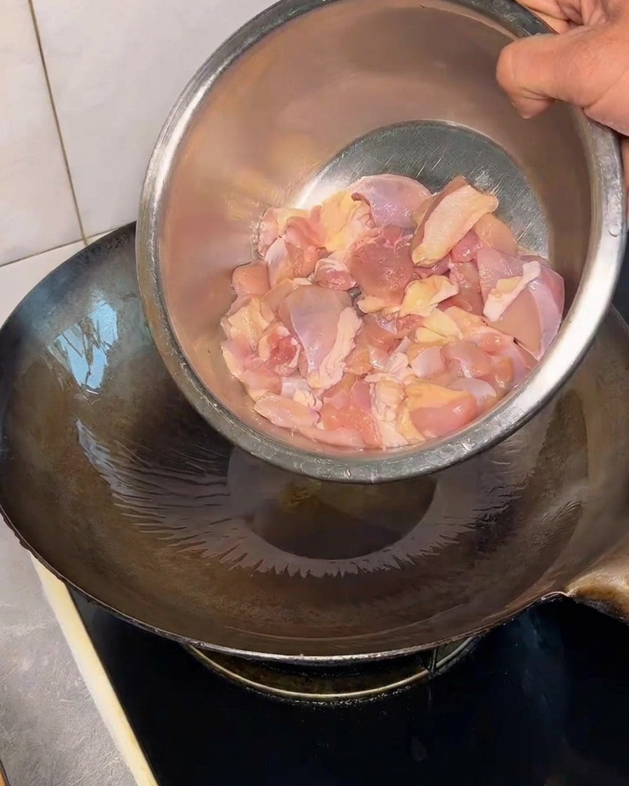 Raw chicken thigh pieces being poured from a metal bowl into a hot, oiled wok.