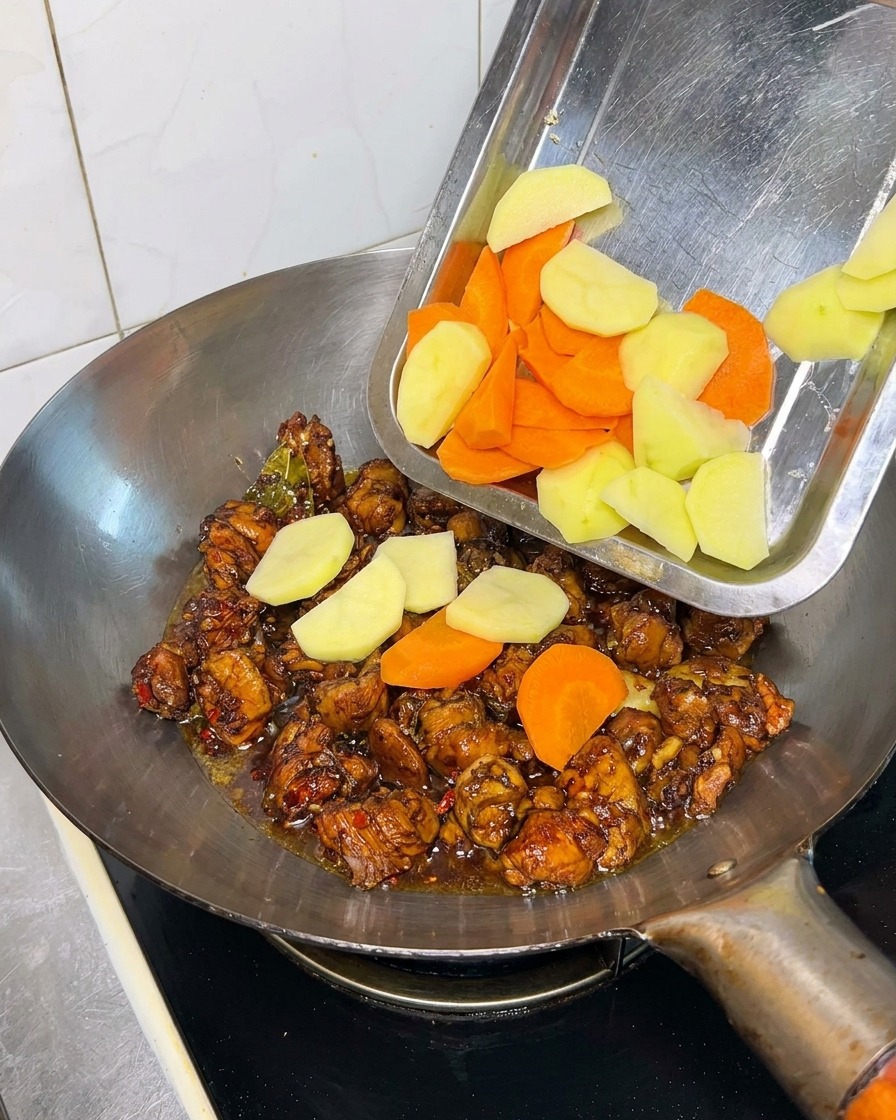 Potato and carrot chunks being poured from a metal tray into a wok containing cooked chicken pieces in a dark sauce.