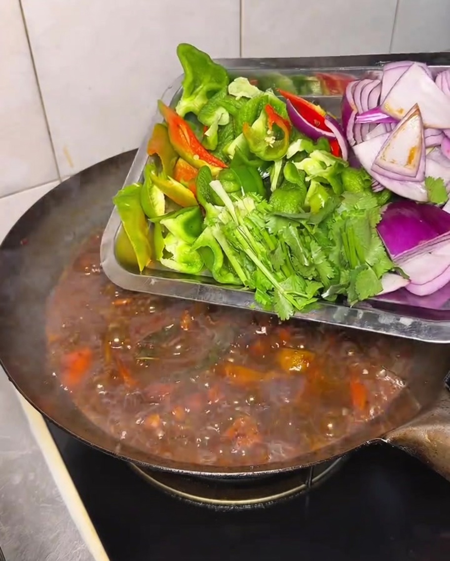 Fresh bell peppers, onions, and cilantro being added to a simmering wok of chicken and vegetable stew.