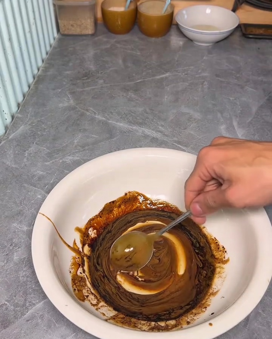 A hand using a spoon to mix a dark, rich sauce consisting of sesame paste and dark soy sauce in a white bowl.