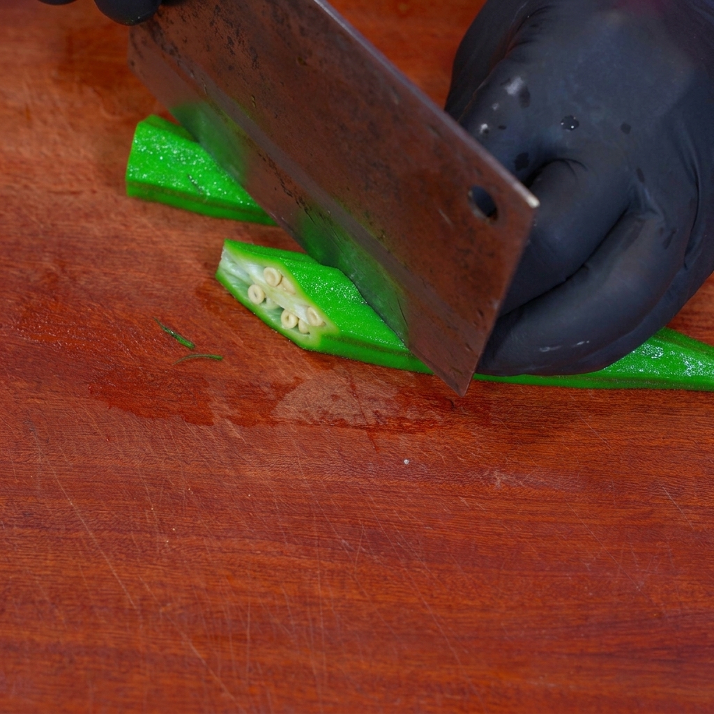 Blanched okra being cut into diamond-shaped pieces on a wooden board.
