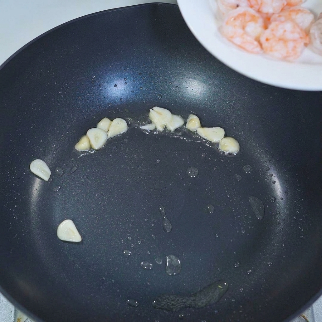 Pre-cooked shrimp being added to a pan with sautéed garlic slices.