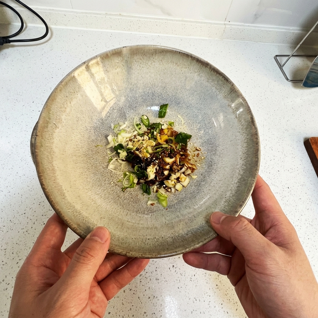A ceramic bowl held by two hands, containing chopped scallions, minced garlic, and liquid seasonings.