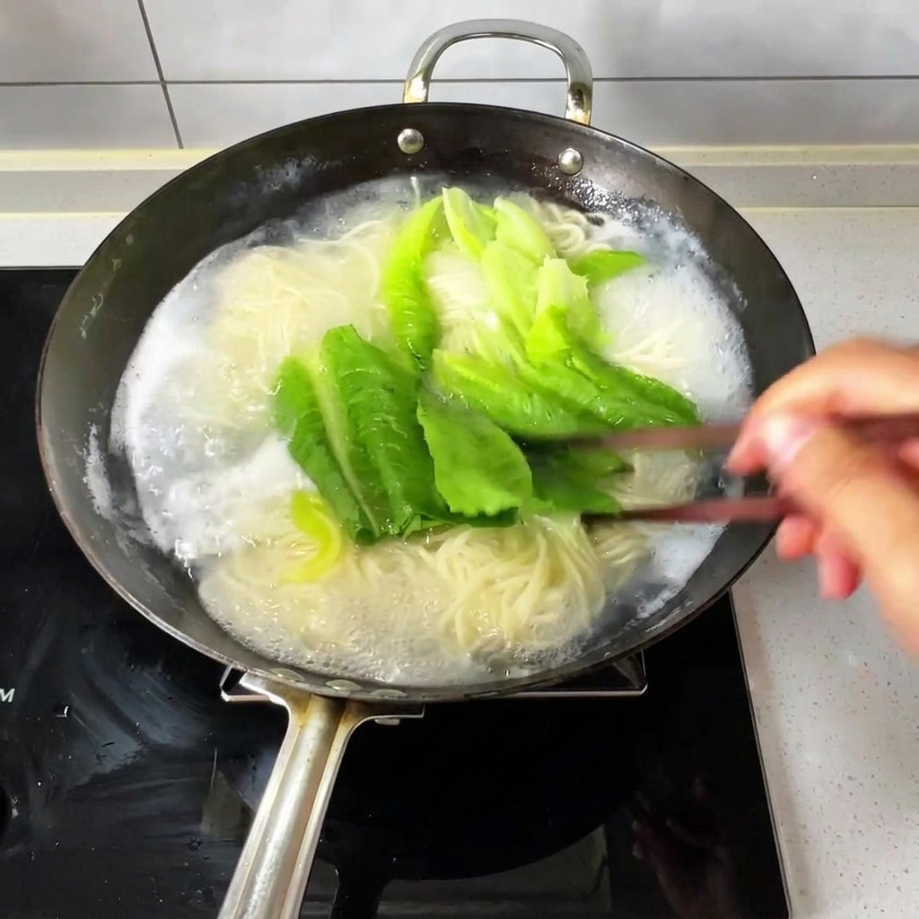 Thin noodles and fresh leafy greens boiling in a wok of bubbling water, being stirred with wooden chopsticks.