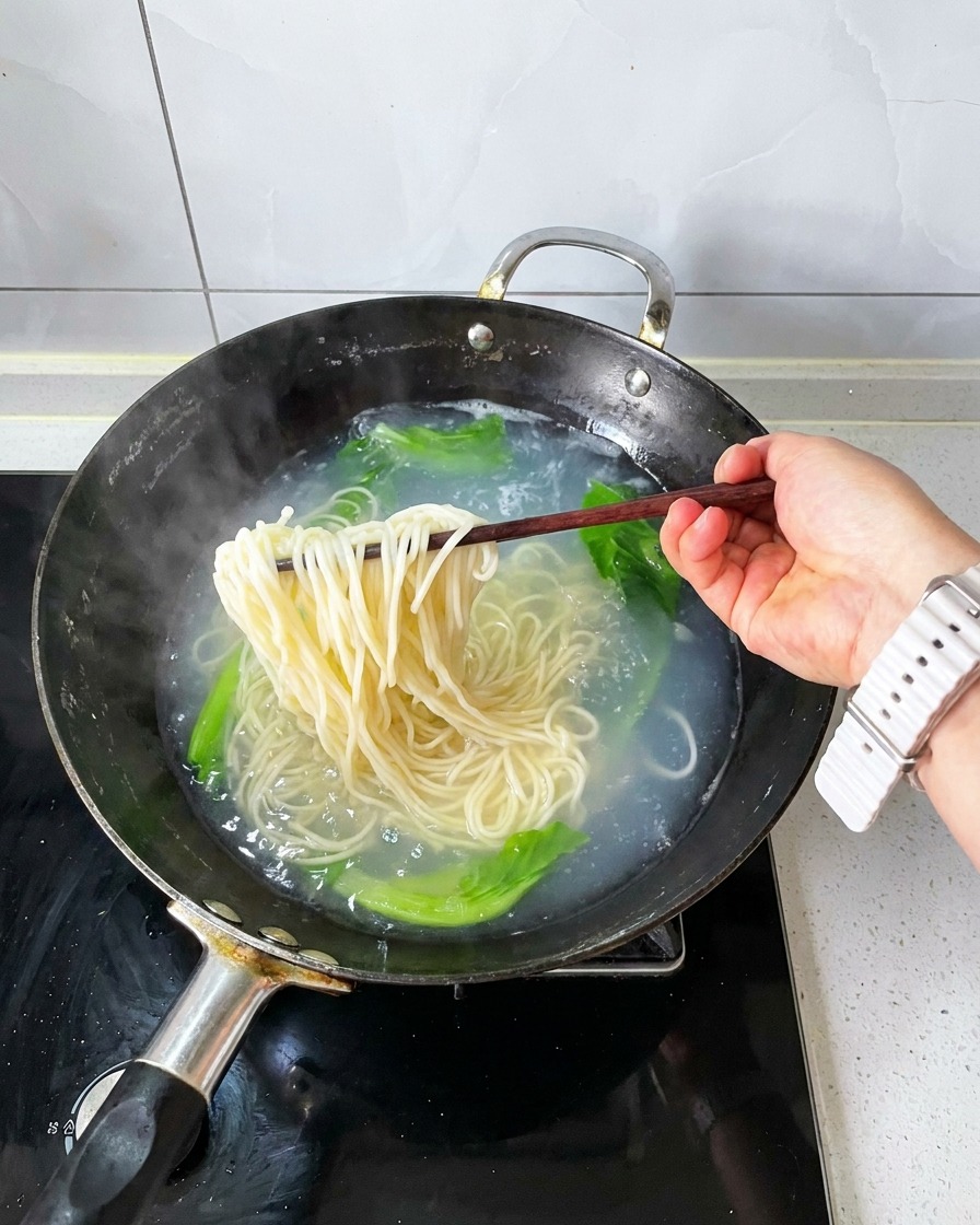 Wooden chopsticks lifting a bunch of cooked noodles and blanched greens out of boiling water.