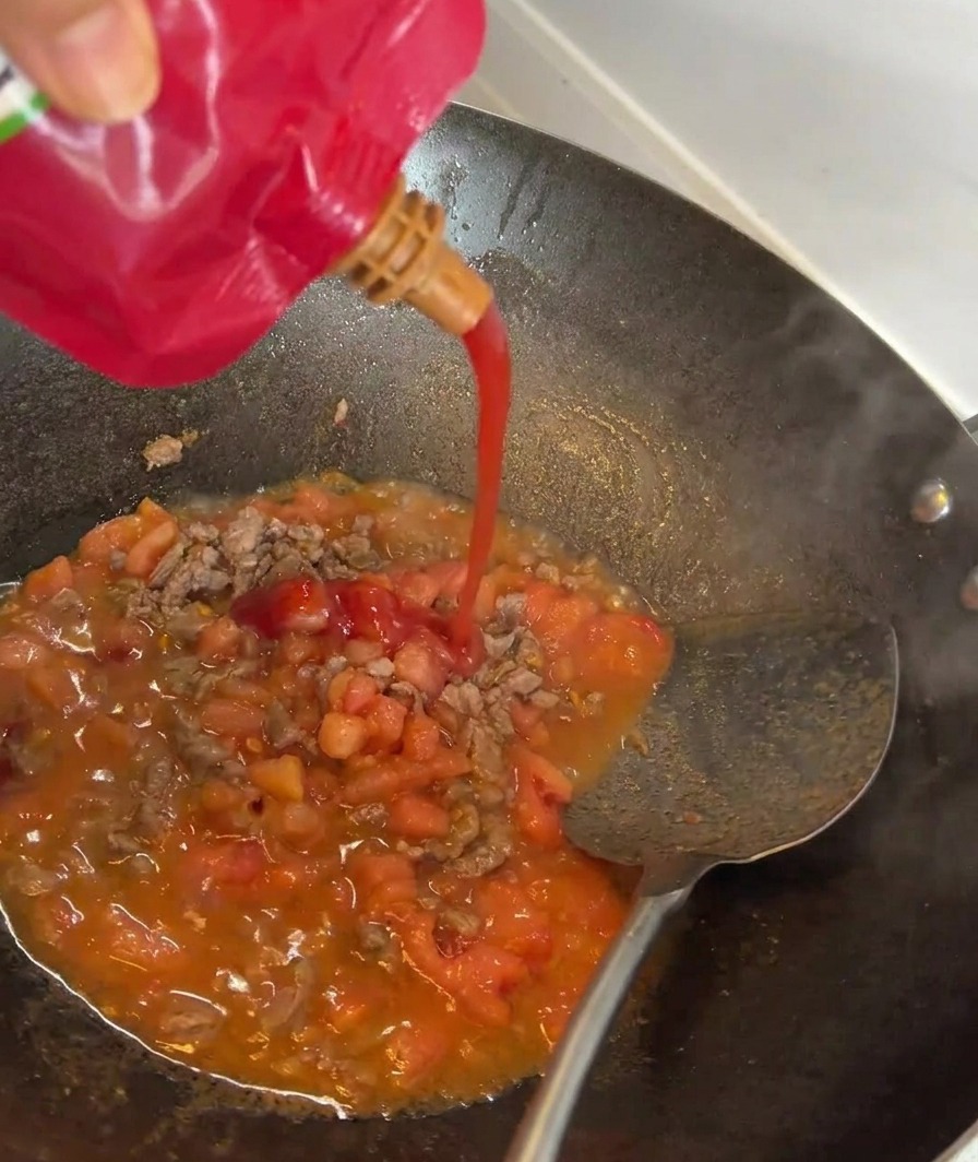 Tomato paste being poured into a pan with sautéed beef and tomatoes.