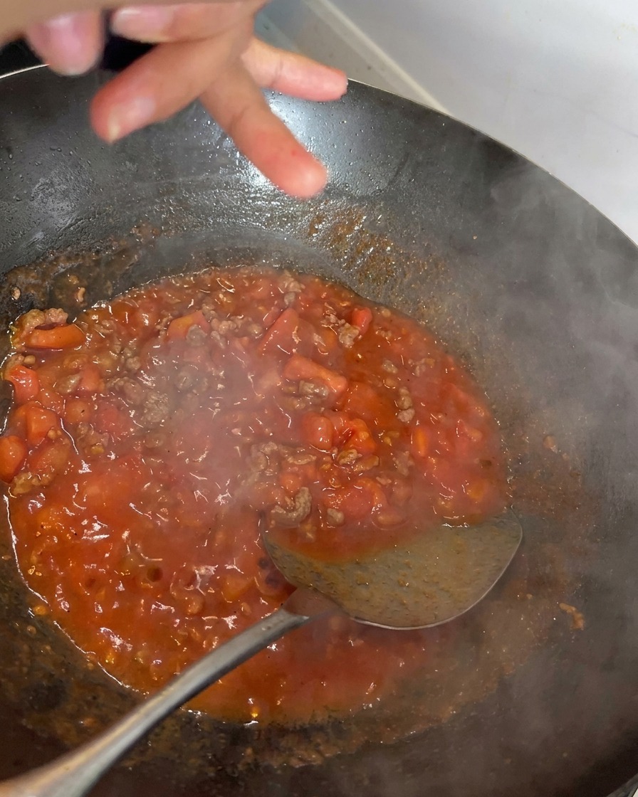 Black pepper being freshly ground over a rich, red simmering tomato and beef sauce in a wok.