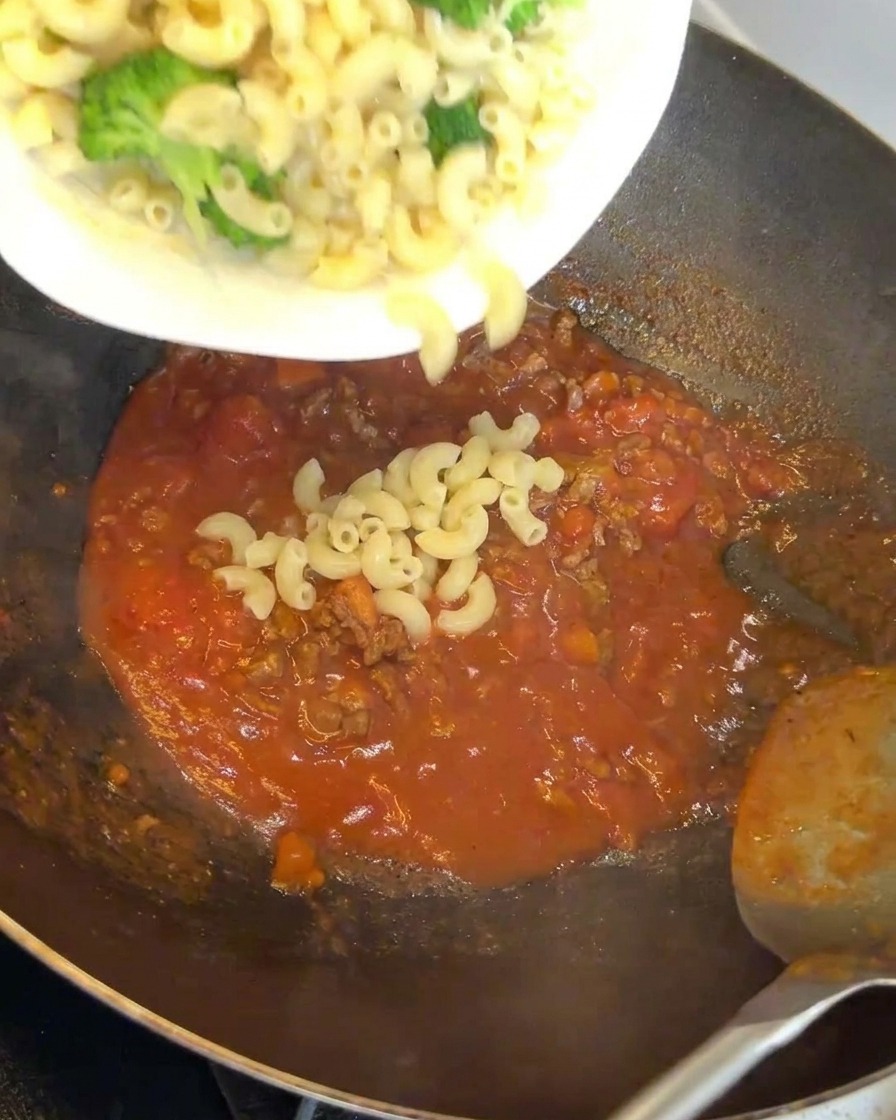 Macaroni and broccoli being added to a wok filled with a red, savory meat sauce.