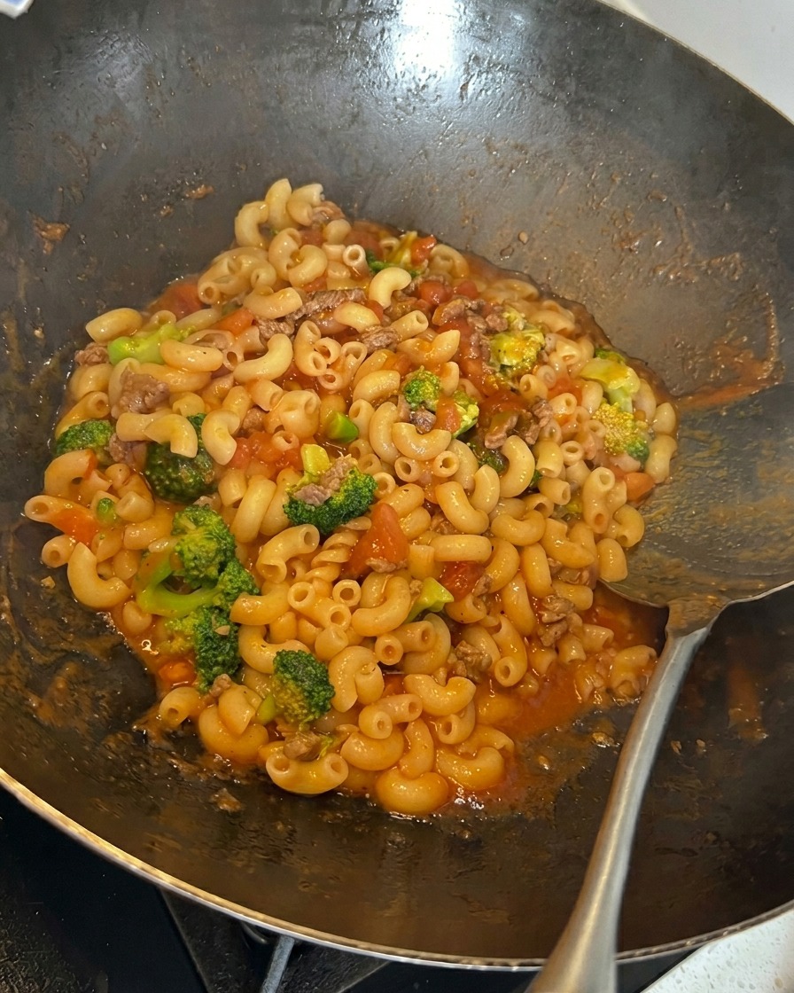 Shredded cheese being sprinkled onto a mixture of macaroni, broccoli, and tomato meat sauce in a wok.