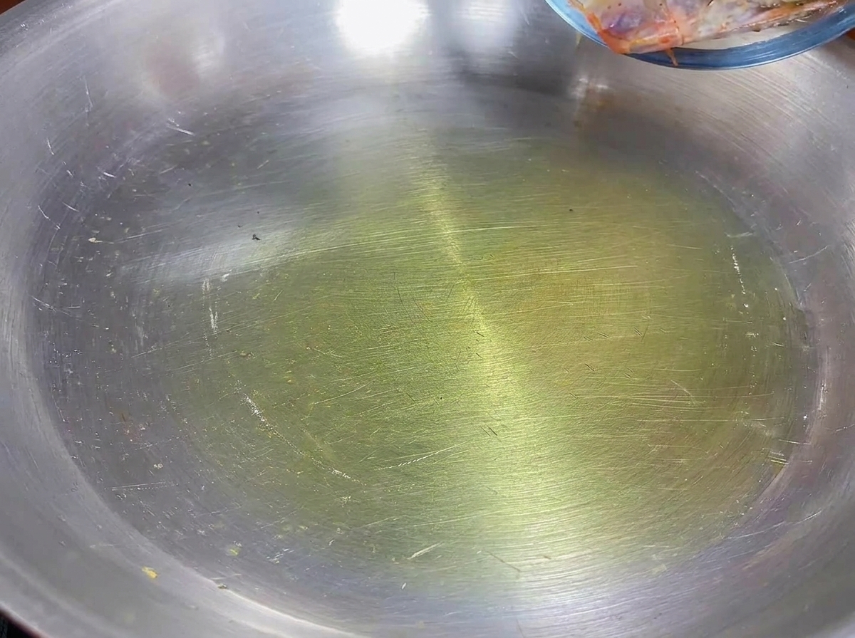 Shrimp heads frying in olive oil in a stainless steel pan.