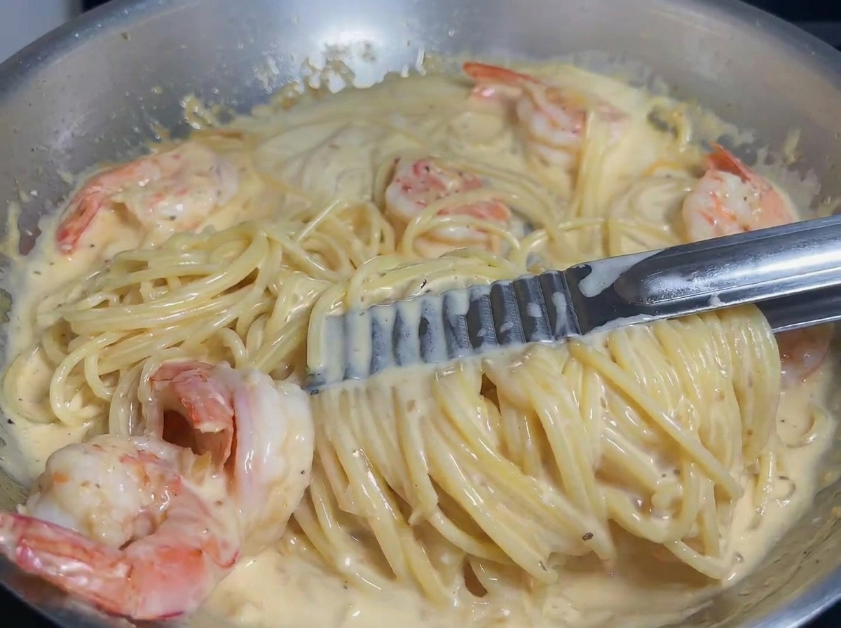 Pasta and shrimp being tossed with tongs in a creamy sauce to ensure even coating.