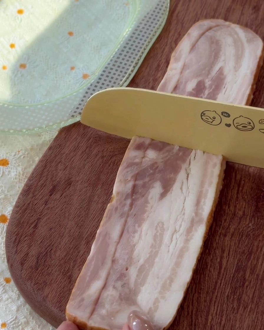 Slices of raw bacon being cut into uniform pieces on a wooden board.