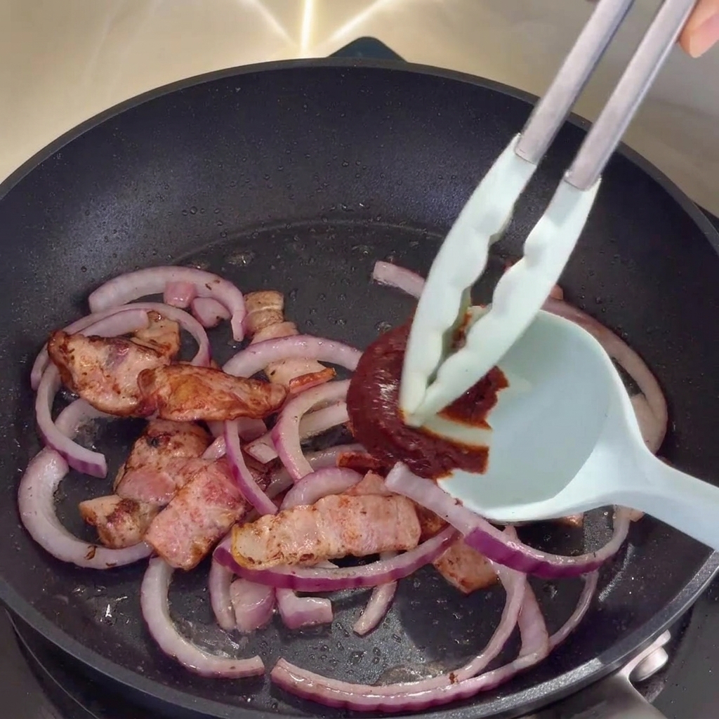 Korean spicy sauce being stirred into the bacon and onion mixture in the pan.