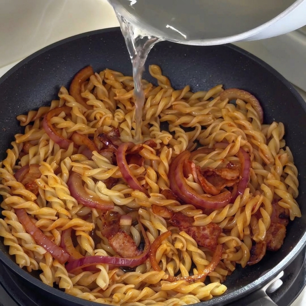 Hot pasta water being poured from a saucepan into a skillet containing fusilli, bacon, and onions.