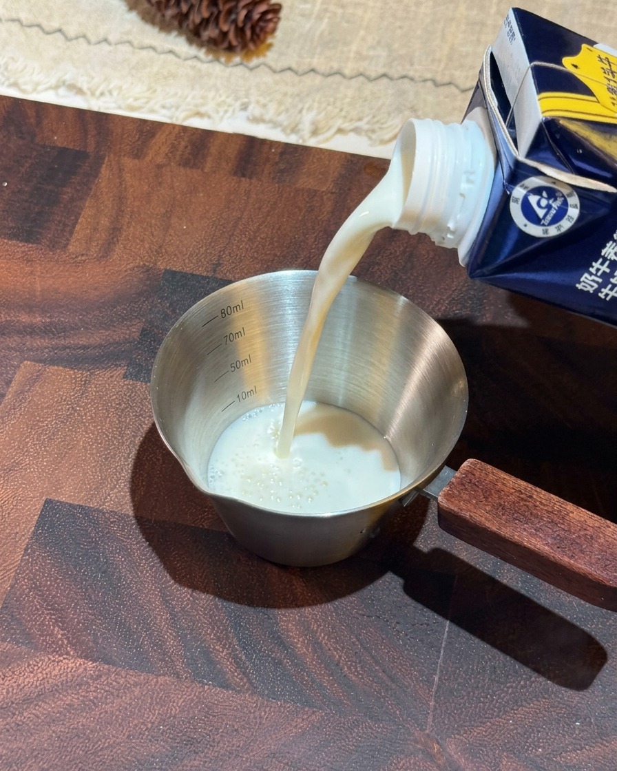 White milk being poured from a blue carton into a small stainless steel measuring cup with a wooden handle.
