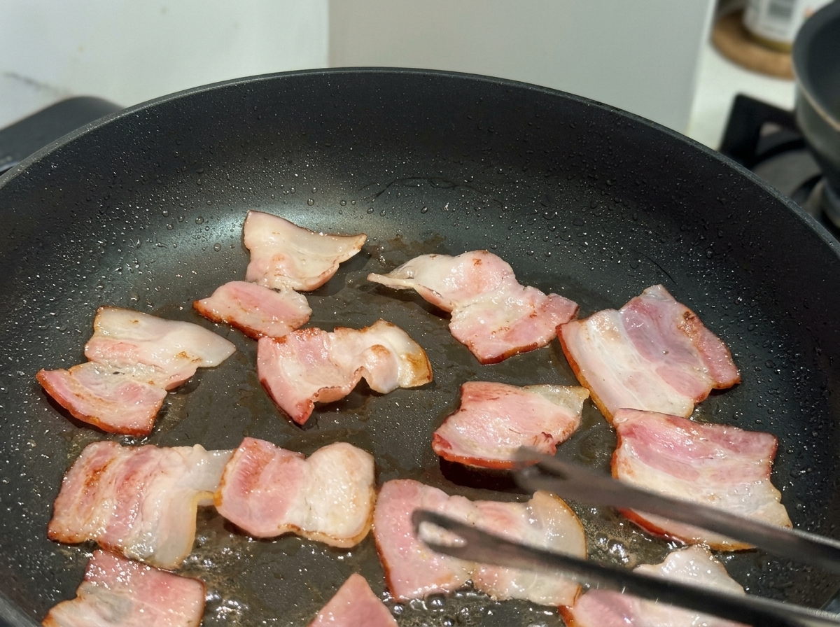 Small, flat pieces of bacon being fried in a black non-stick pan with a pair of tongs visible.