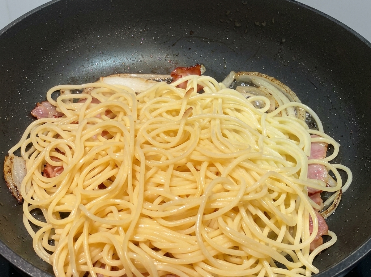 Cooked spaghetti noodles piled on top of sautéed onions and pieces of bacon in a black pan.