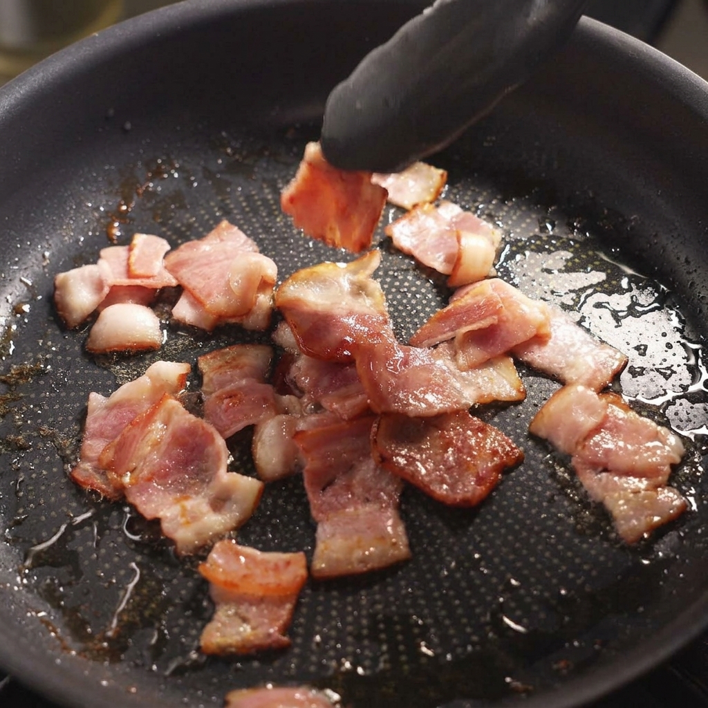 Black tongs flipping pieces of slightly browned, sizzling bacon in a skillet.
