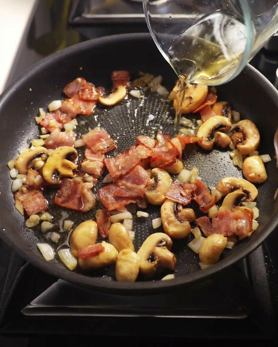 Clear white wine being poured from a glass measuring cup into a pan with cooked mushrooms and bacon.