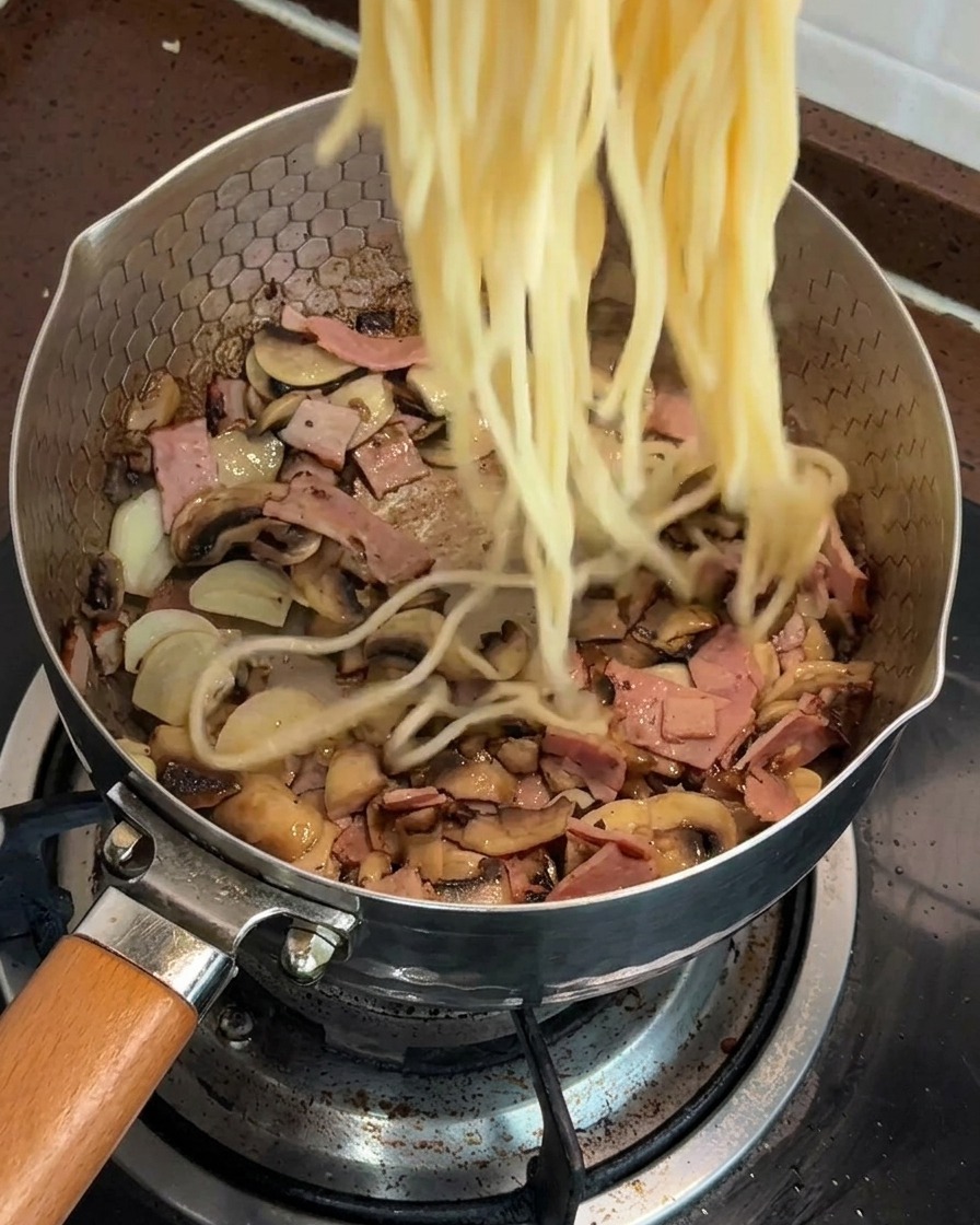 Cooked spaghetti being added to a pan filled with a mixture of sautéed mushrooms and bacon.