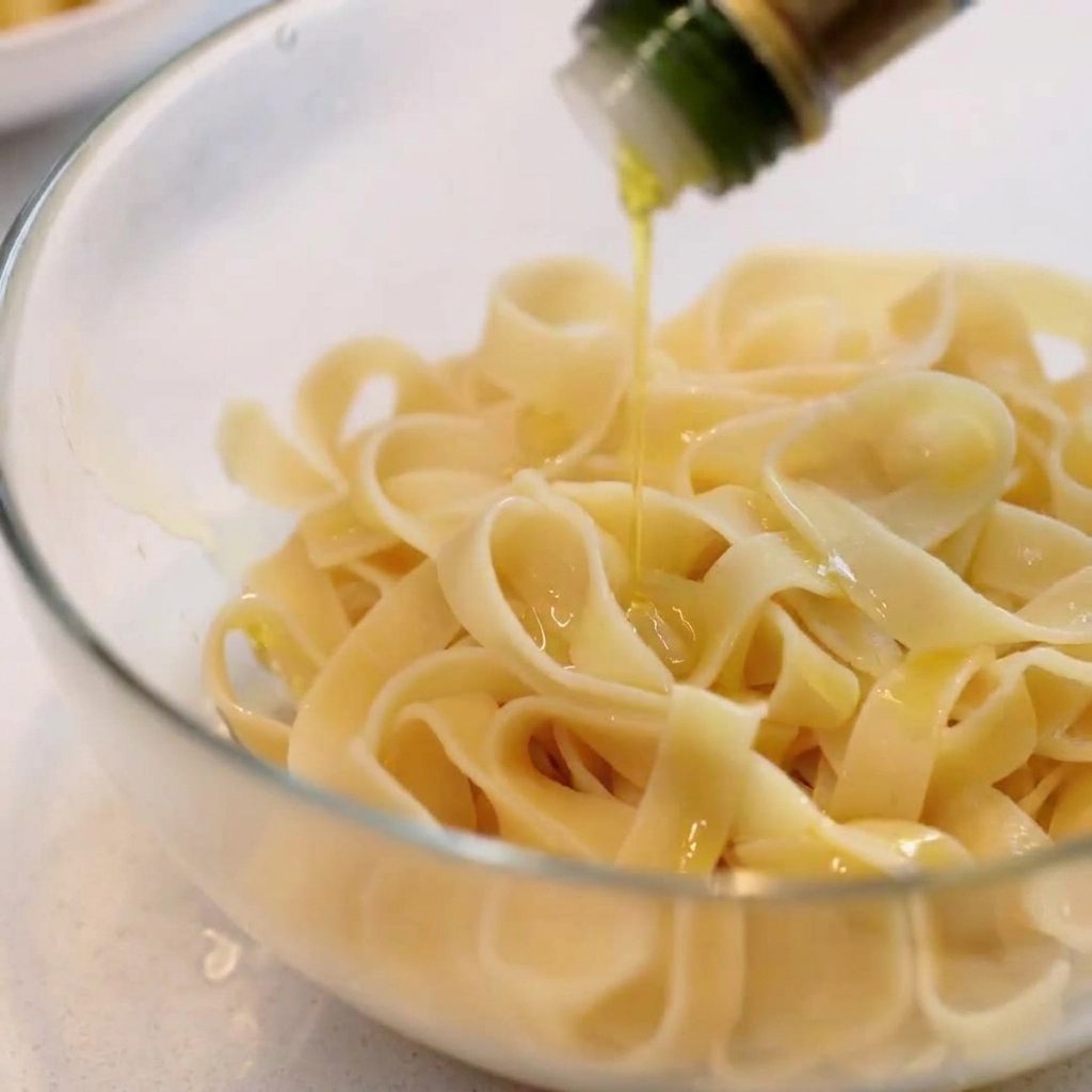 Cooked wide pasta in a clear glass bowl being drizzled with olive oil.