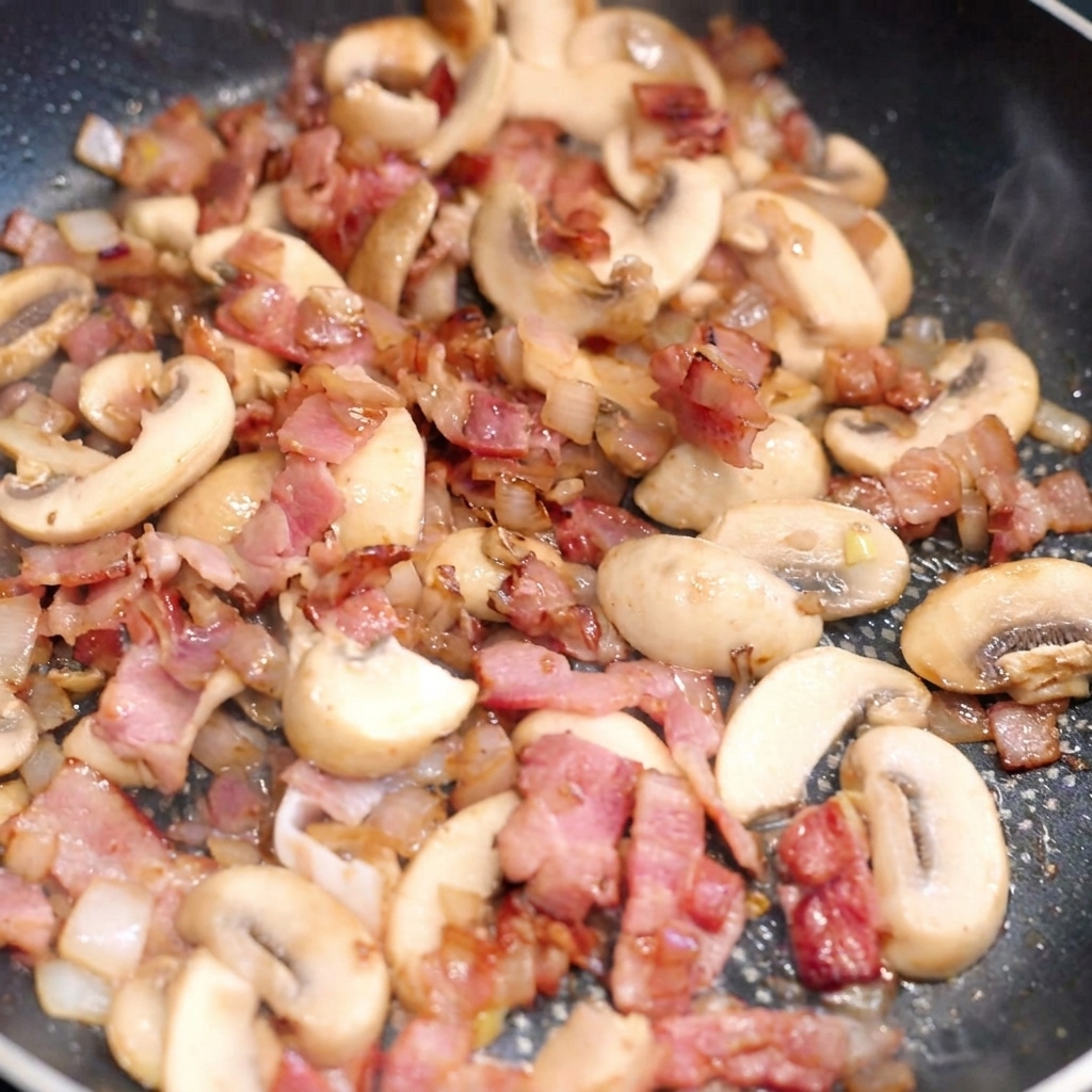 Wide pasta being mixed into a pan filled with browned bacon, sautéed mushrooms, and onions.