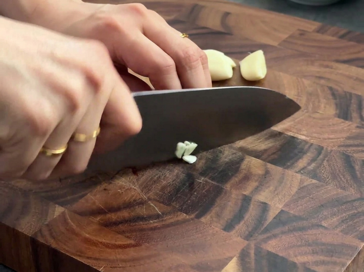 Cloves of fresh garlic being minced on a wooden cutting board with a chefs knife.