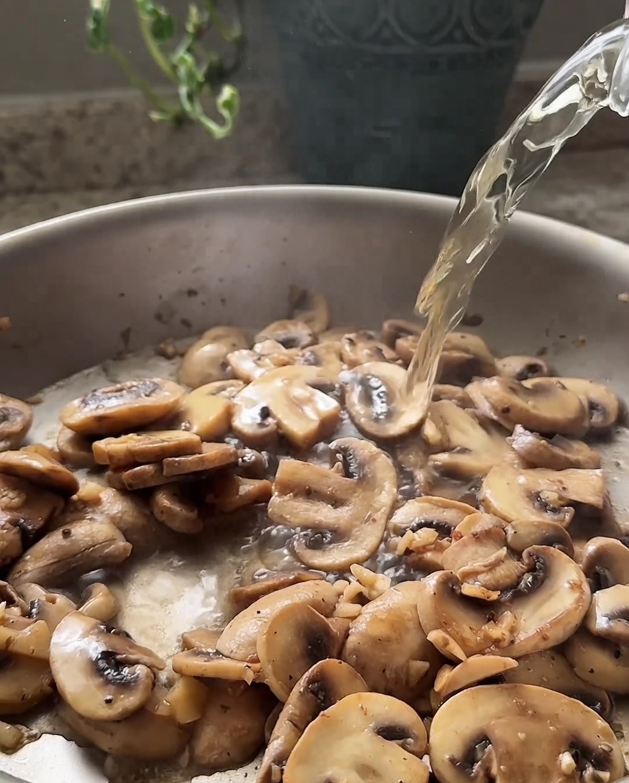 White wine being poured from a glass into a pan containing sautéed sliced mushrooms.