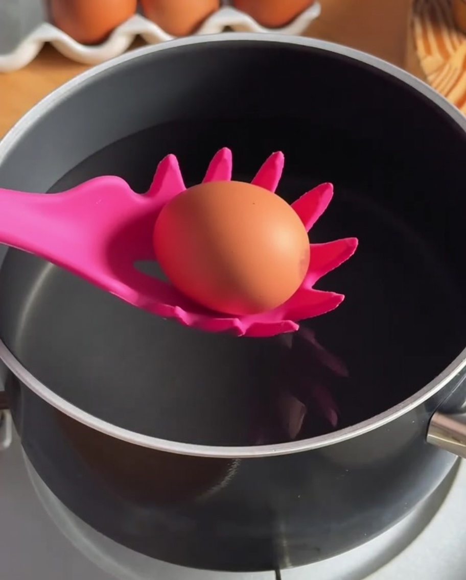 An egg being lowered into a pot of hot water using a silicone slotted spoon.