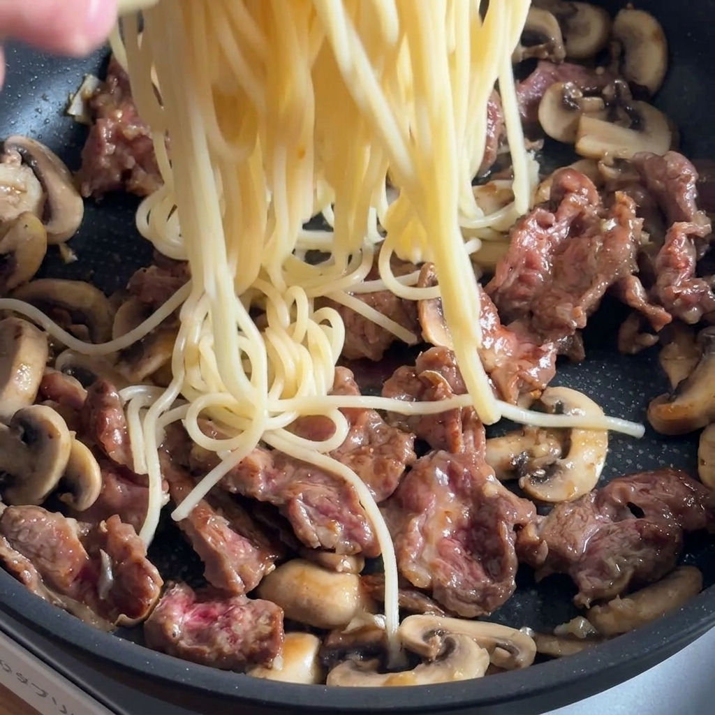 Pasta being added to a pan with beef and mushrooms, ready for seasoning.