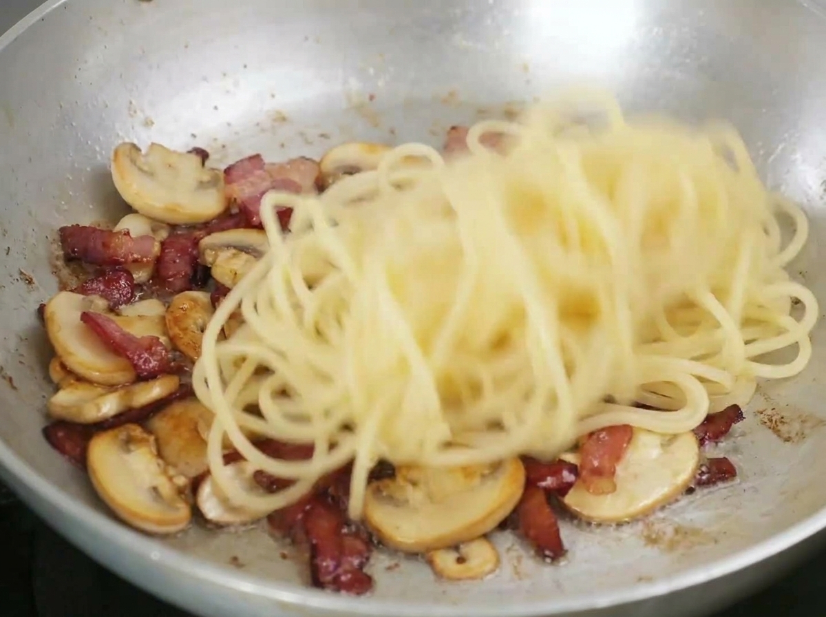 Spaghetti being tossed in a pan with fried bacon and sliced mushrooms.