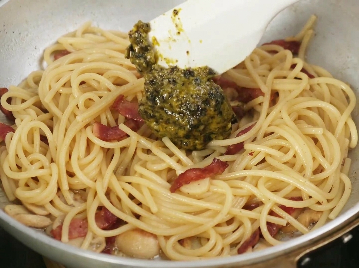 A spatula mixing dark, aromatic black truffle sauce into a pan of cooked pasta, garlic, and bacon.
