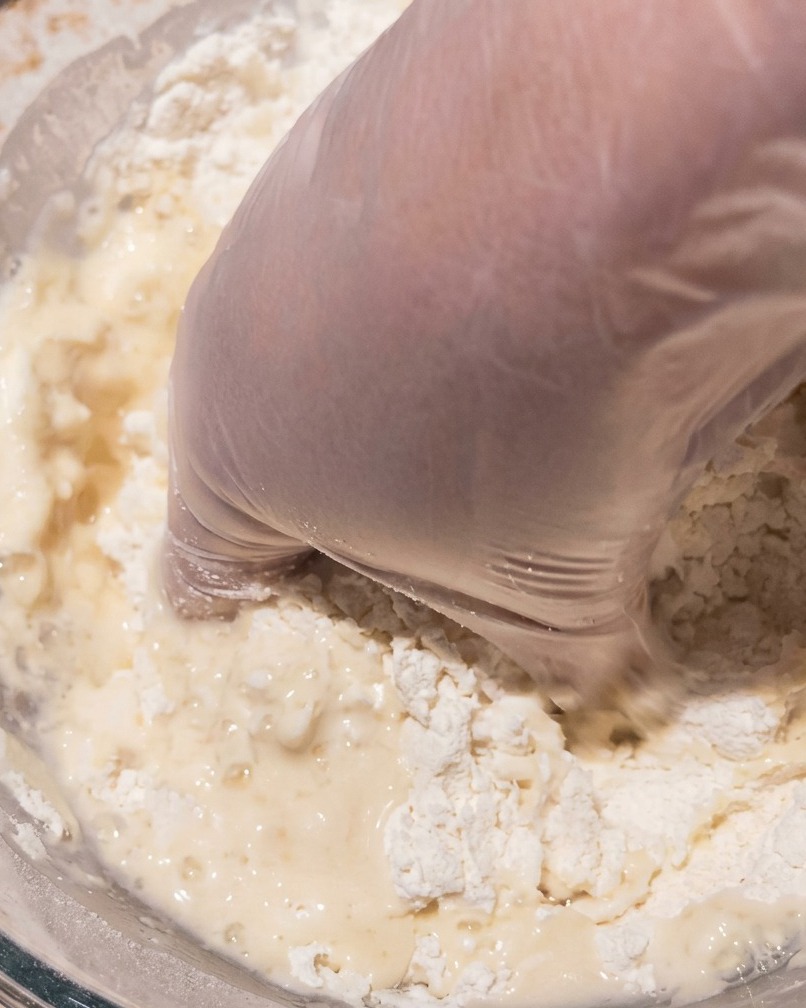 A gloved hand mixing sticky, wet dough in a large glass bowl.