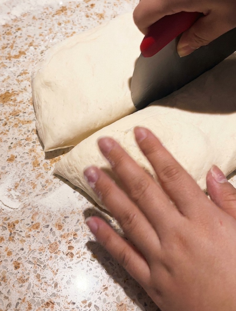 A metal bench scraper cutting into a piece of fermented pizza dough on a countertop.
