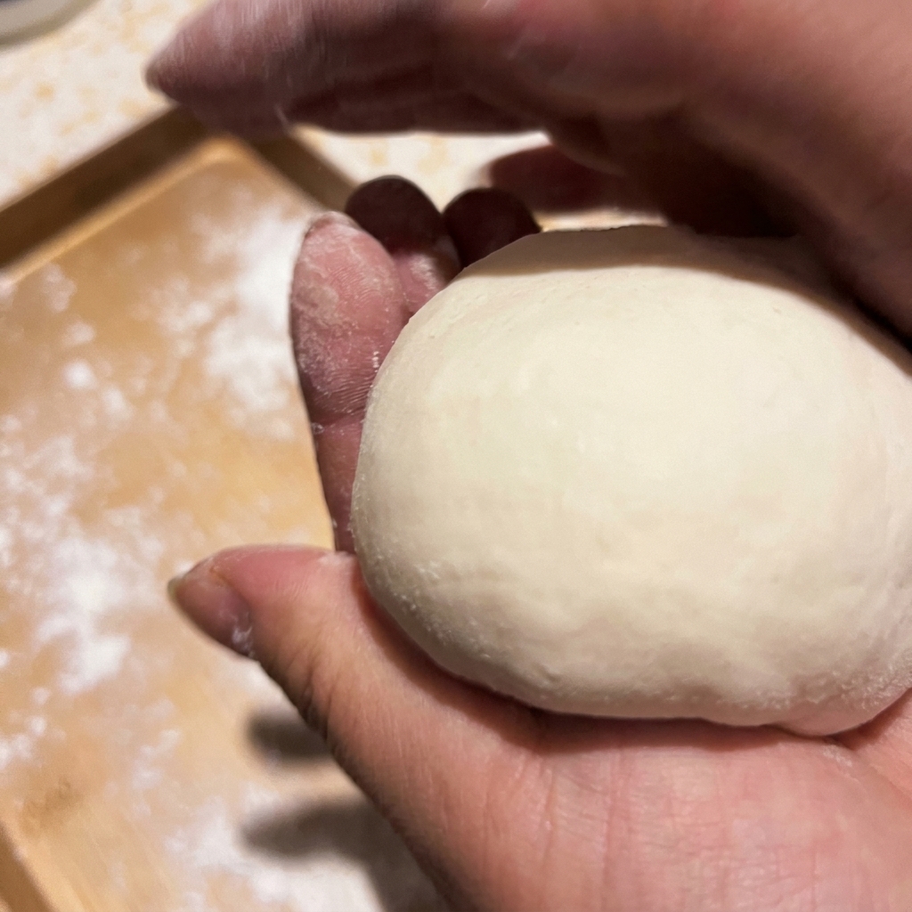 Hands cupping and shaping a portion of raw pizza dough into a smooth ball.