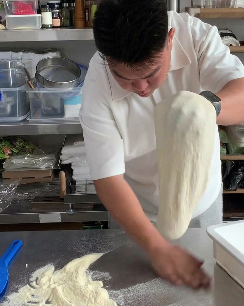 Hands shifting the stretched pizza dough back and forth to dust off excess flour.