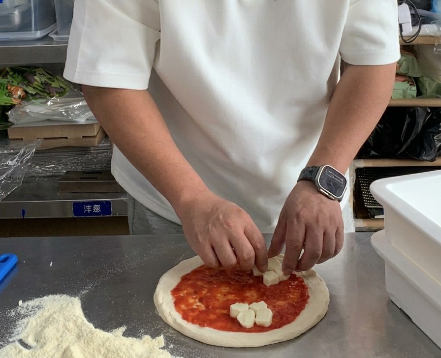 Hands placing cubes of fresh mozzarella onto a tomato sauce-covered pizza dough.