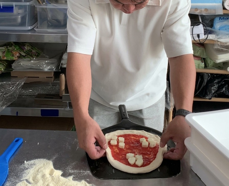 Hands adjusting the round edges of an unbaked Margherita pizza resting on a perforated metal pizza peel.