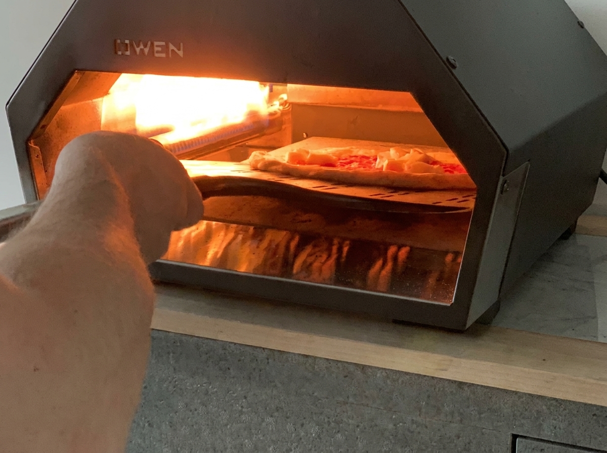 A pizza peel sliding an unbaked Margherita pizza into a hot tabletop gas pizza oven.