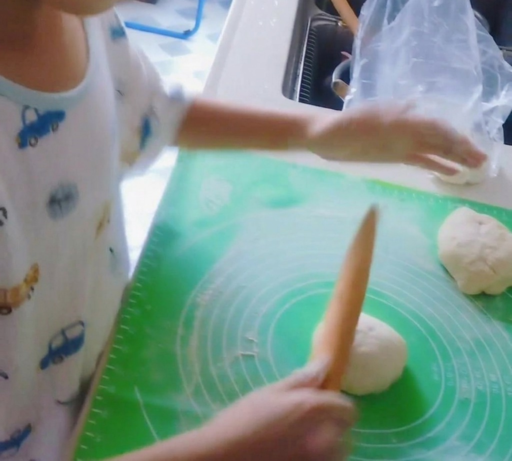 A wooden rolling pin flattening a ball of pizza dough into a circular crust.
