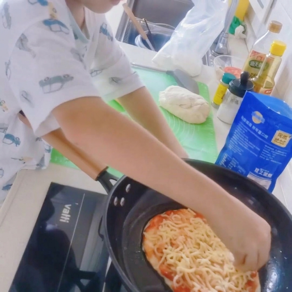 A close-up of a pizza base in a frying pan being topped with a thick layer of shredded mozzarella cheese.