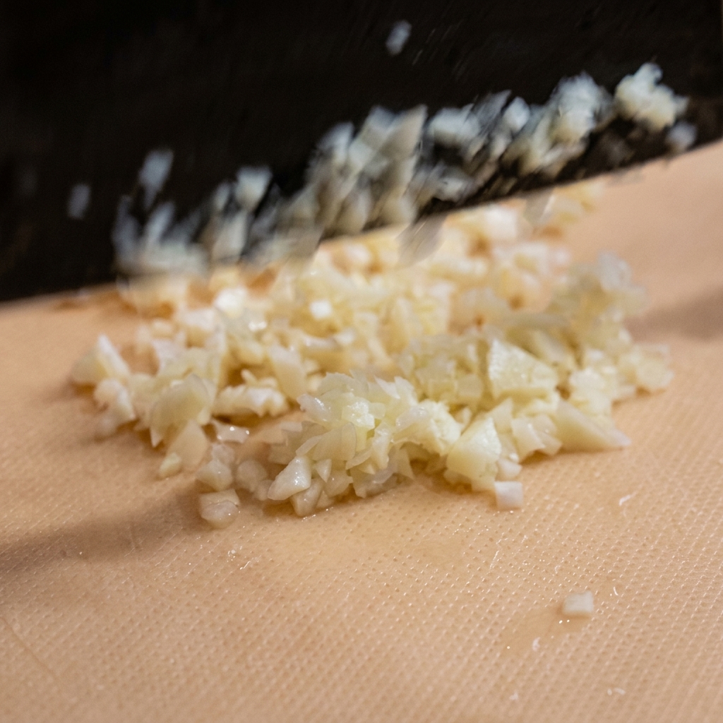 Finely minced garlic pieces scattered on a light-colored cutting board being prepared for the recipe.