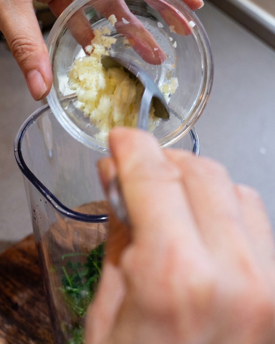 A blender container being filled with minced garlic, freshly grated cheese, and toasted pine nuts.