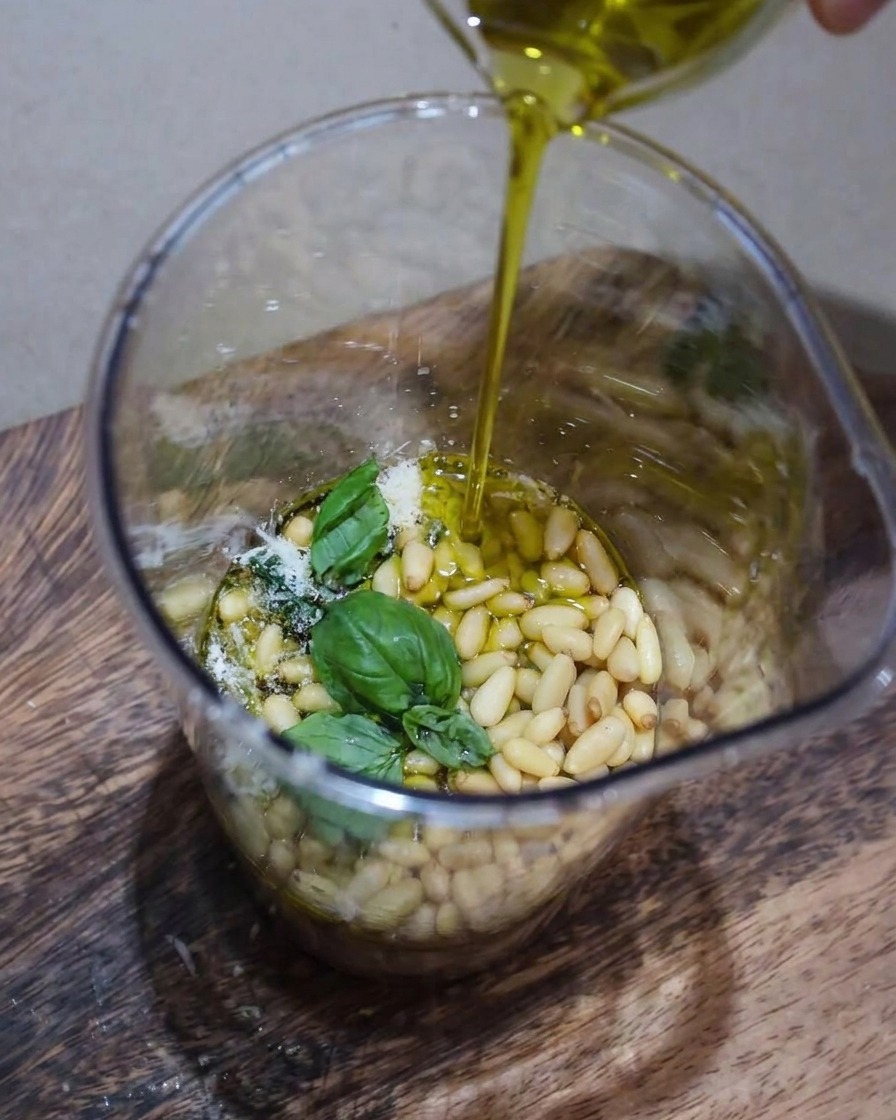 A close-up view of pesto ingredients including fresh basil, pine nuts, and Parmesan being blended in a container with a thin stream of olive oil pouring in.