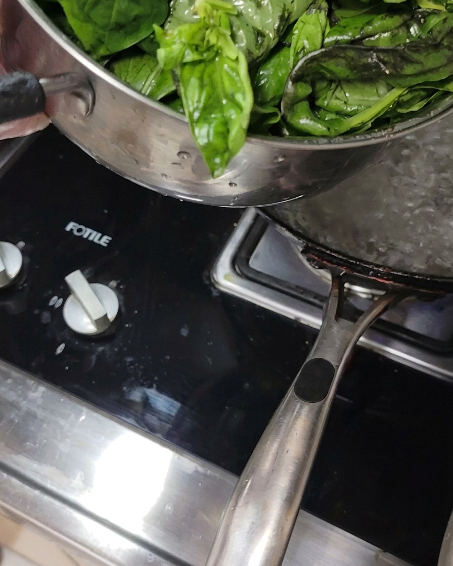Fresh basil leaves being submerged in a pot of boiling water on a stove.