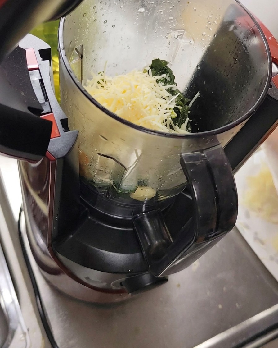 Ingredients including grated cheese, garlic, and pine nuts placed into a food processor with basil leaves.