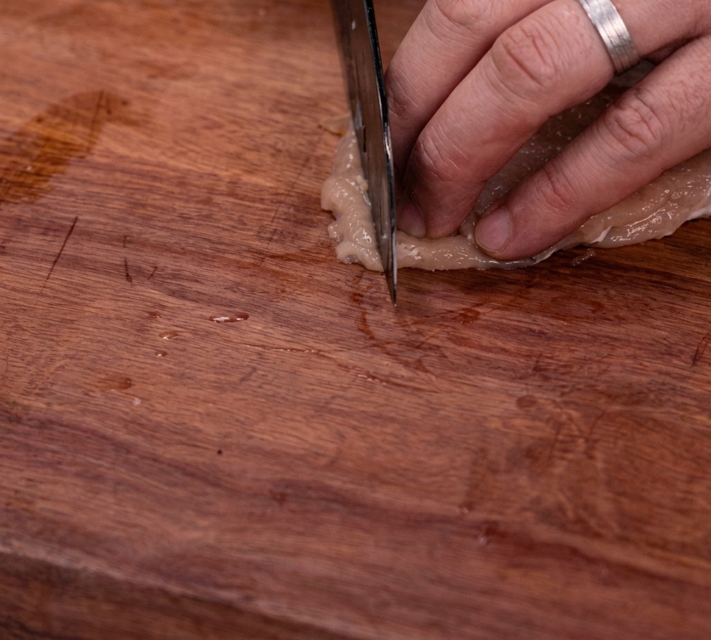 Raw chicken breast being cut into uniform strips on a wooden cutting board.